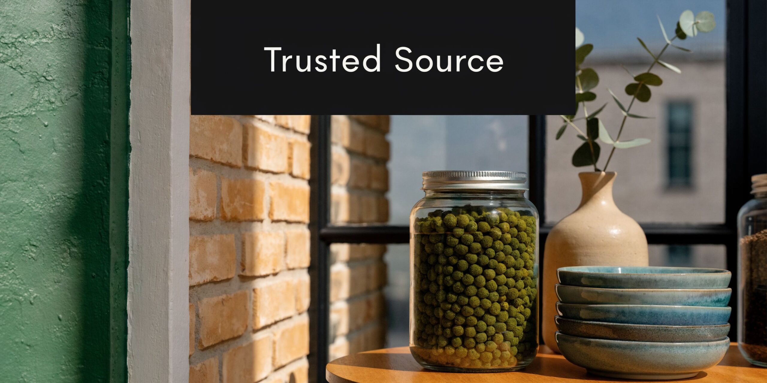 A glass jar filled with small green herbal pellets displayed on a wooden surface with decorative bowls.